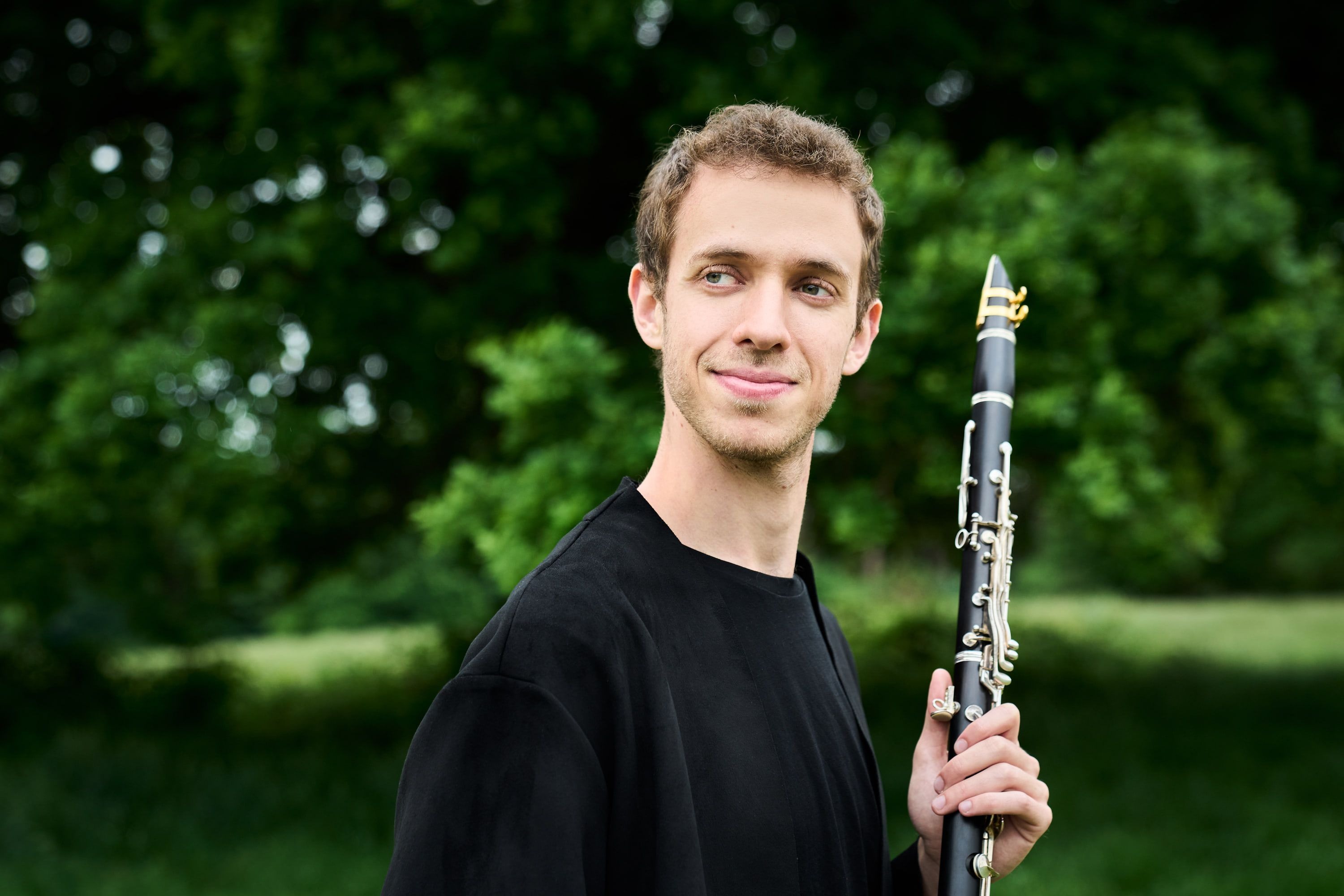 Jonathan Leibovitz smiling over his shoulder in a green park, clarinet raised