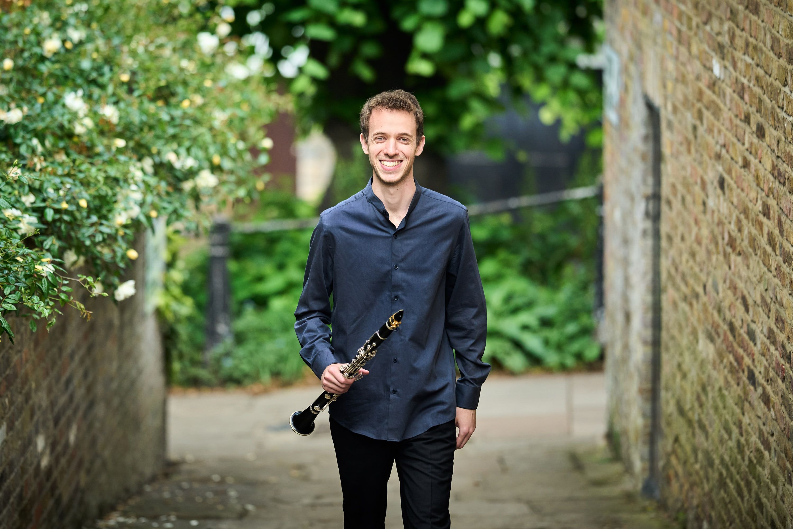 Jonathan Leibovitz holding his clarinet on a garden path between brick walls